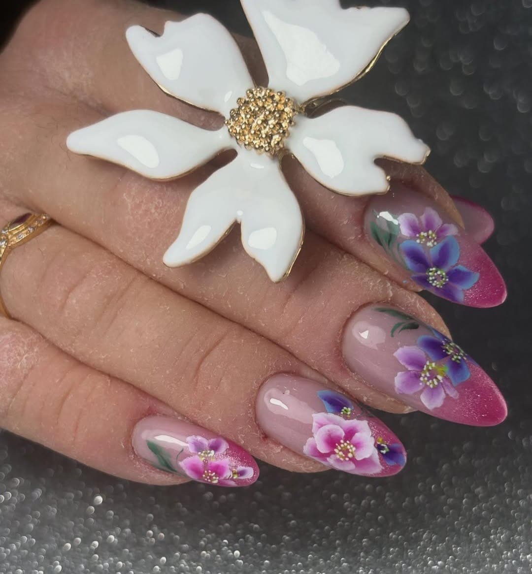 Unhas em amêndoa com base nude, pintura floral em roxo, azul e rosa e pontas em bordô