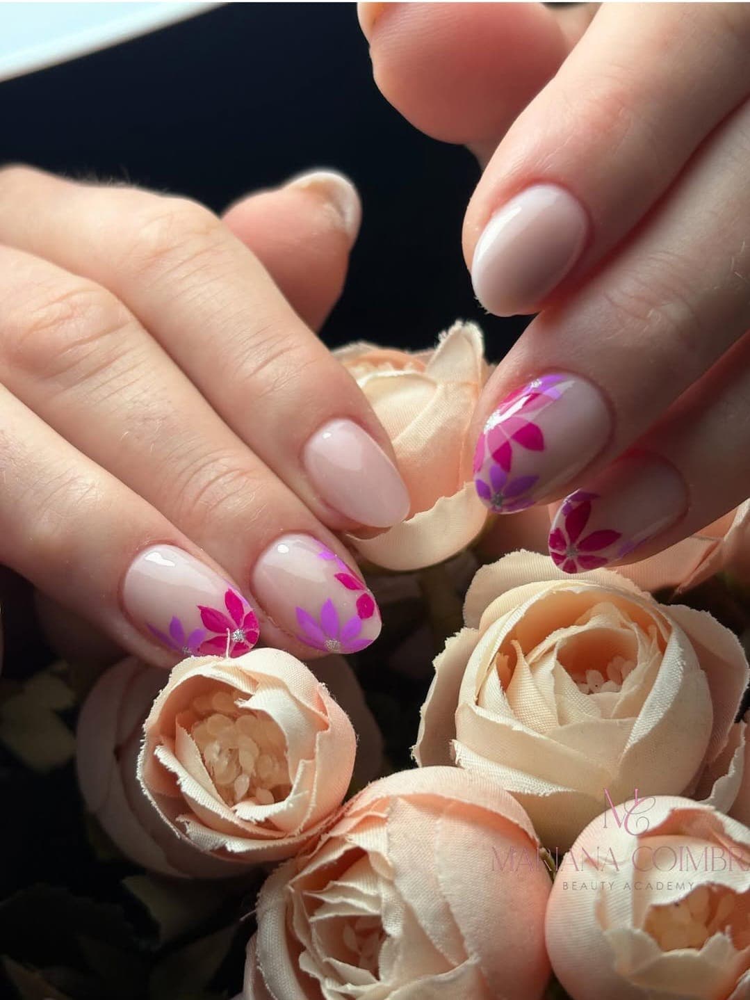Unhas ovais curtas com base nude e flores pintadas em tons de rosa e roxo